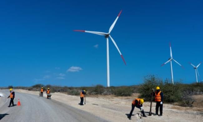 Six men in h-vis jackets working along a road, with three turbines looming over them