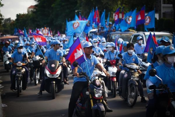 A crowd of people on motorbikes, dressed in pale blue and waving flags, drive down a tree-lined street with cars