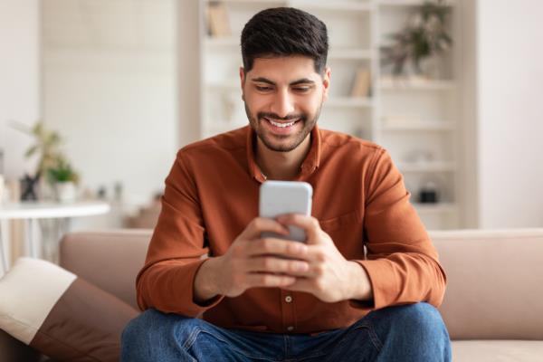 young man smiling and looking up information on his cell phone while sitting on his couch