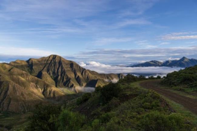 A view of a dirt road hugging a hillside with rugged green peaks above clouds