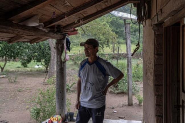 A man stands on the veranda of a wooden house with a garden behind him 
