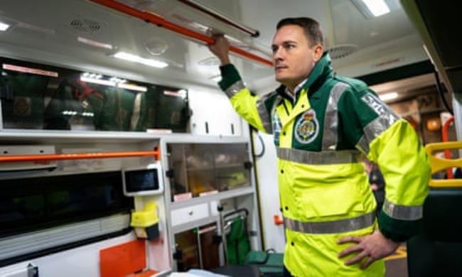 Wes Streeting dressed in  paramedic’s hi-vis green and yellow jacket, standing in the back of an ambulance
