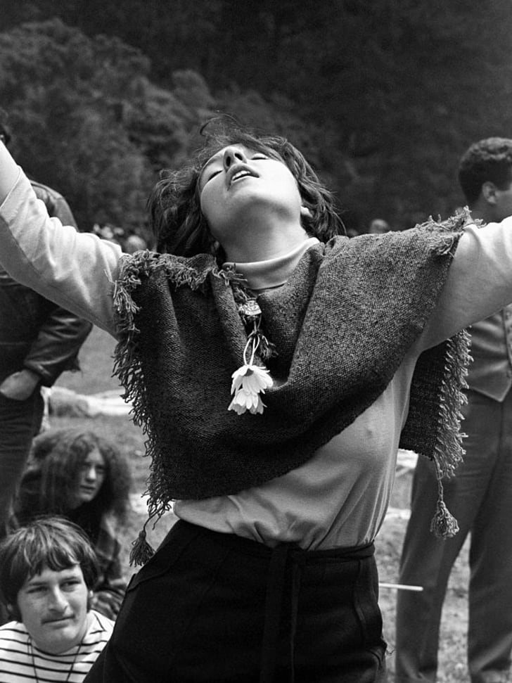 black and white photo of a scarf-wearing woman dancing, arms tossed up in ecstasy, as people in the background look on