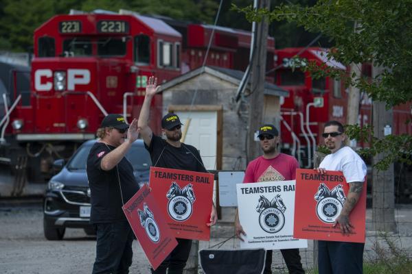 A group of men in t-shirts and ba<em></em>seball caps holding signs that say 'Teamsters Rail' stand in front of a statio<em></em>nary freight train