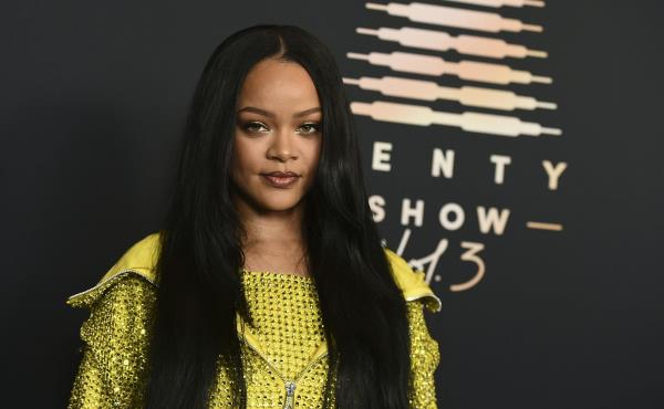 A young Black woman with long, black hair and a glittery yellow top poses in front of a backdrop that says 'Savage X Fenty Show Vol. 3'