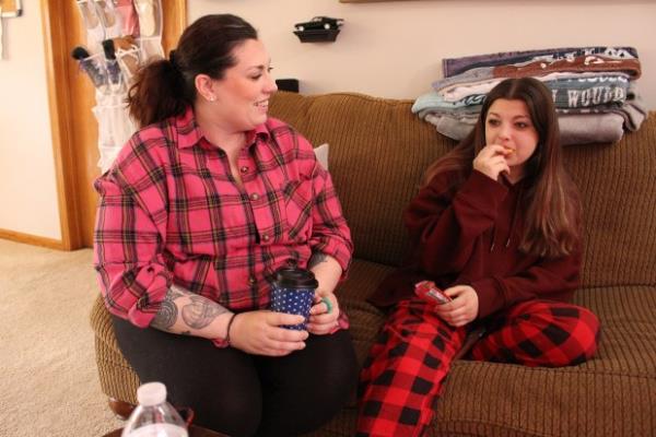 A woman in a red plaid shirt sits on a couch talking to her daughter, who is wearing red plaid pajama pants